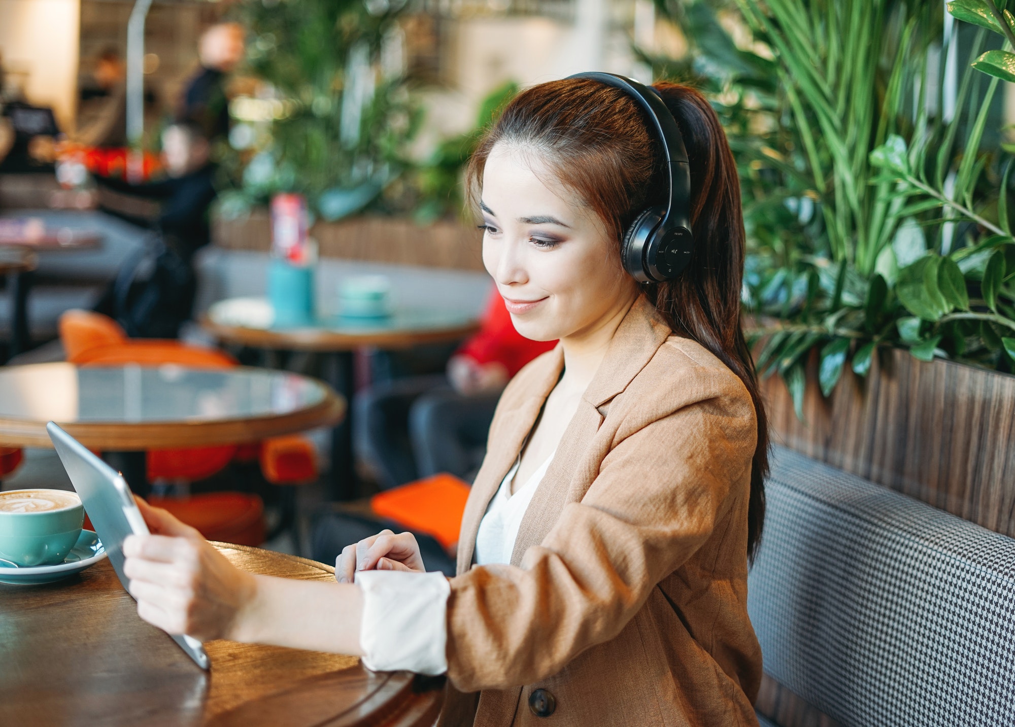 Young smiling asian girl student in headphones communicates by tablet at cafe