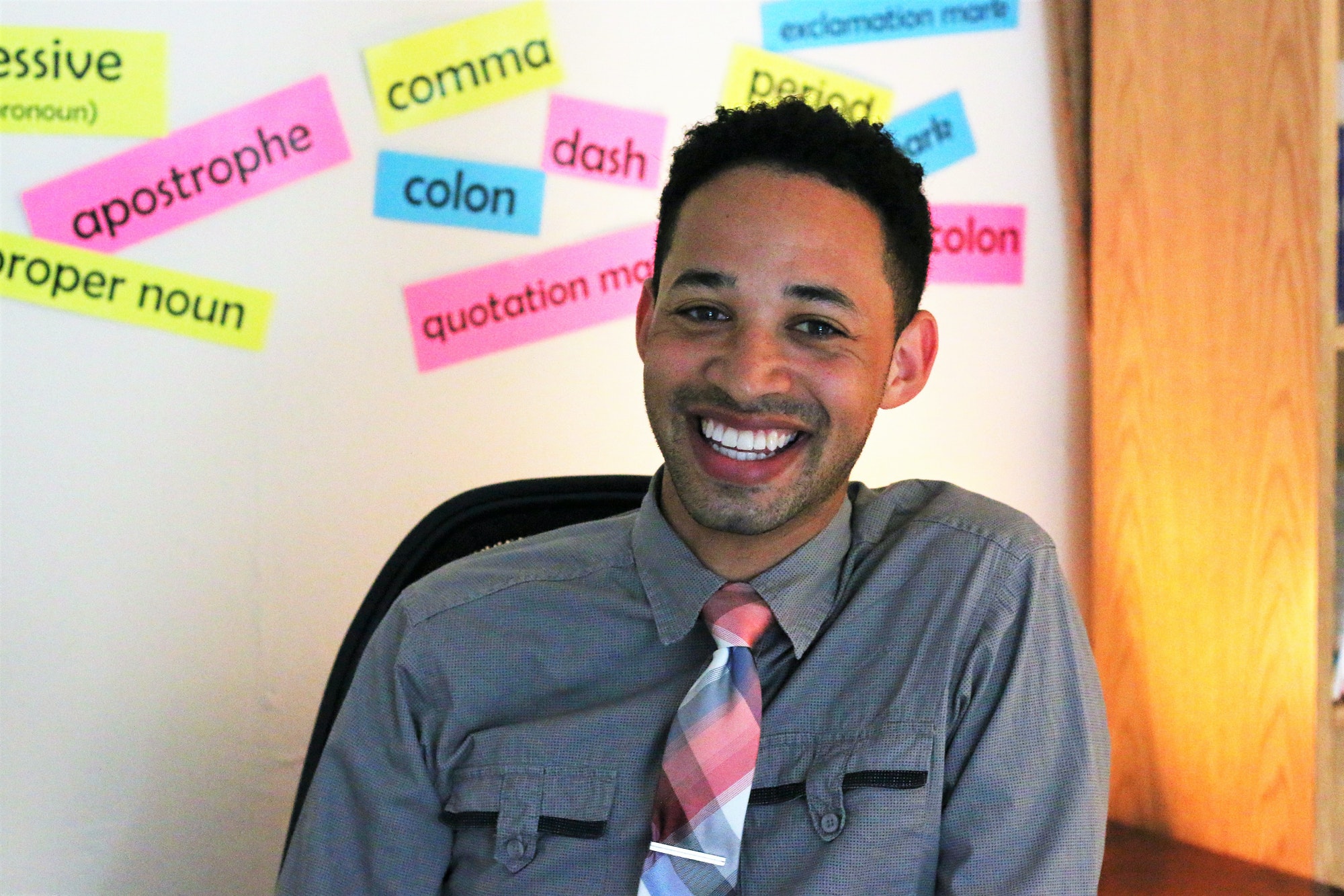 A young diverse teacher sitting in his classroom in front of a board with vocabulary words