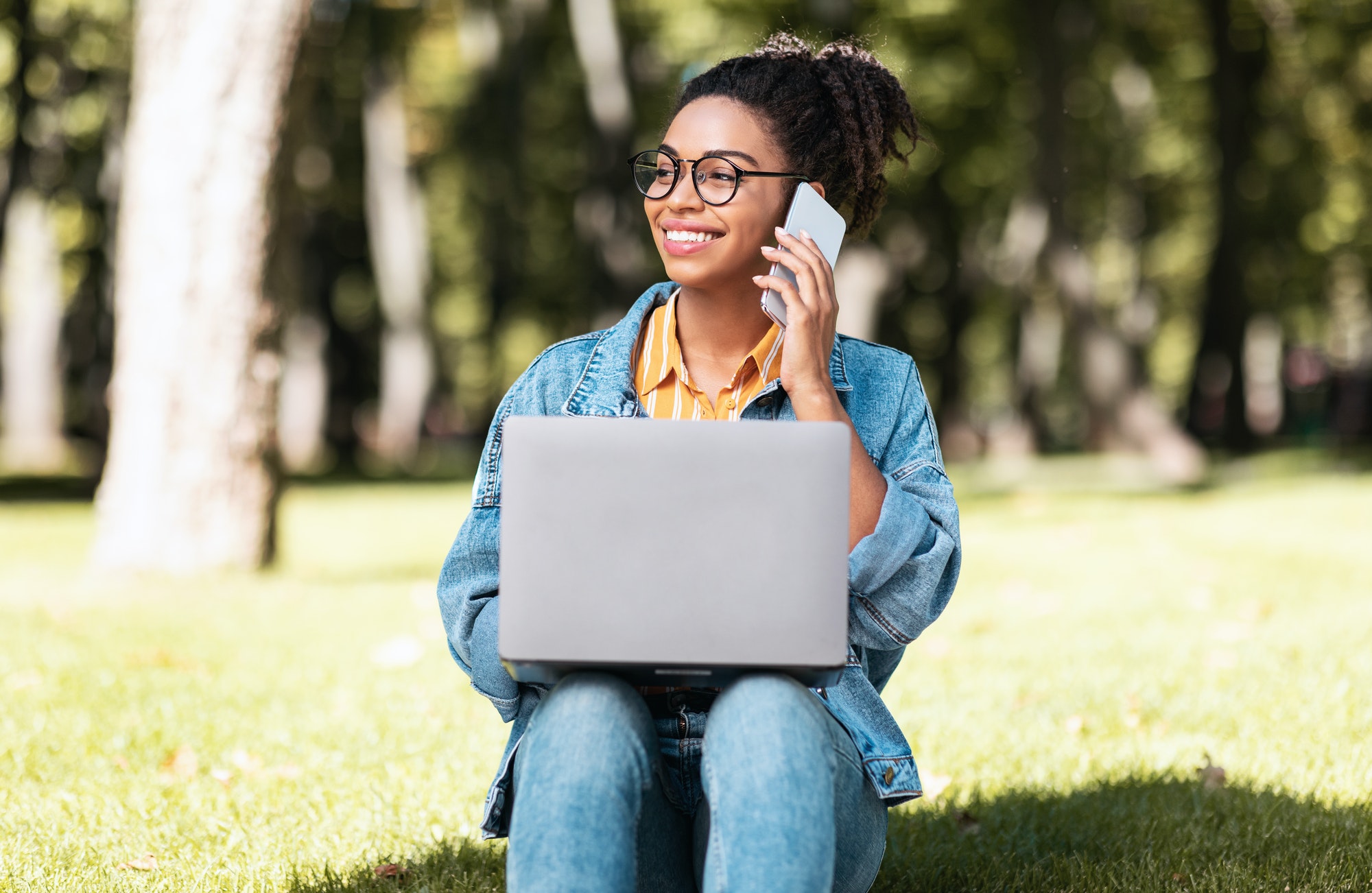 Smiling Black Student Girl Speaking On Cellphone Using Laptop Outdoors