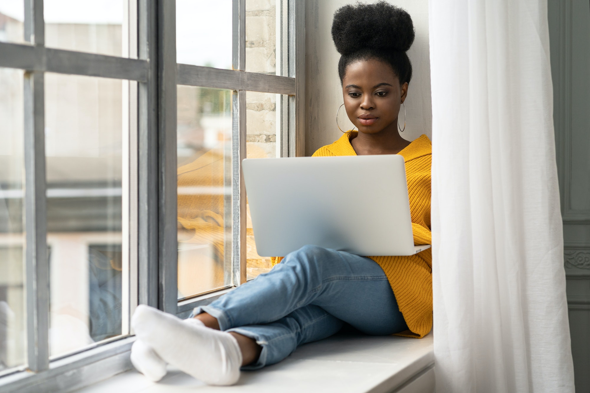 Black woman sitting on windowsill, working on laptop, distance online learning preparing for an exam